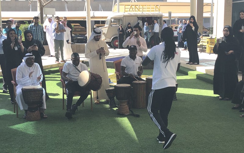 A group playing djembes together in a circle outdoors, with the facilitator leading
