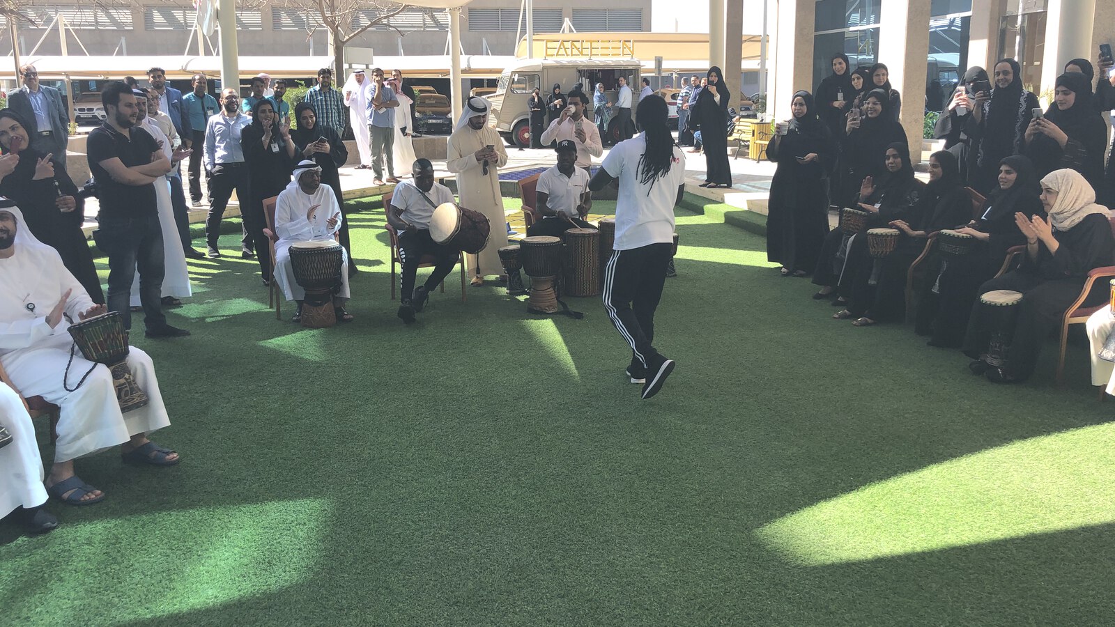 A group playing djembes together in a circle outdoors, with the facilitator leading