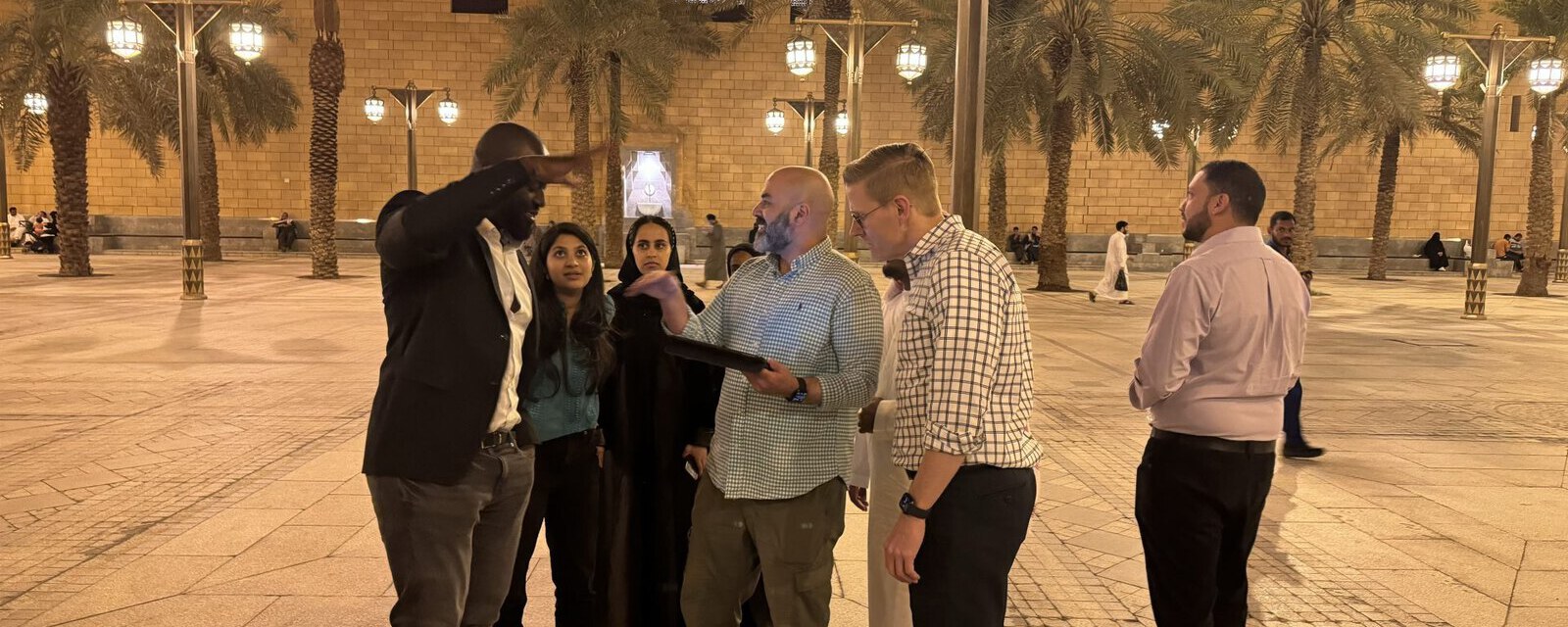A team gathered around a tablet in a Riyadh square at dusk